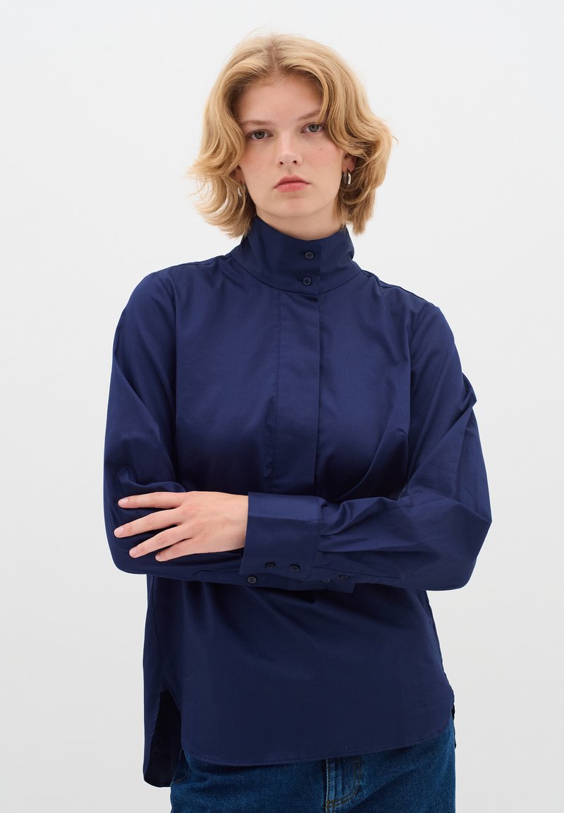 Camicia color blu navy con colletto alto, chiusura frontale con bottoni e maniche lunghe. Il tessuto appare liscio, con spacchi laterali all'orlo.