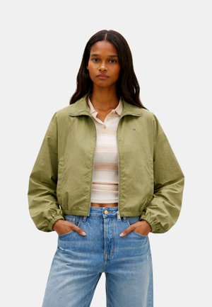 Donna che indossa una giacca verde oliva chiaro con cerniera sopra una polo beige a righe e jeans blu a vita alta, in piedi con le mani nelle tasche.