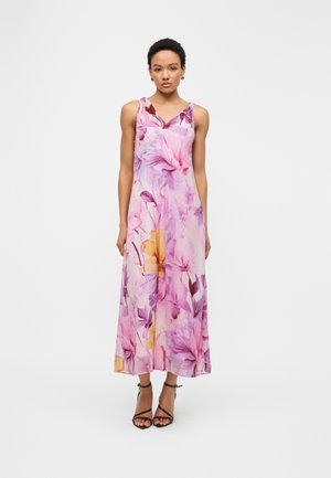 Donna in piedi che indossa un abito lungo senza maniche rosa con fiori e tacchi neri con cinturini, su uno sfondo bianco uniforme.