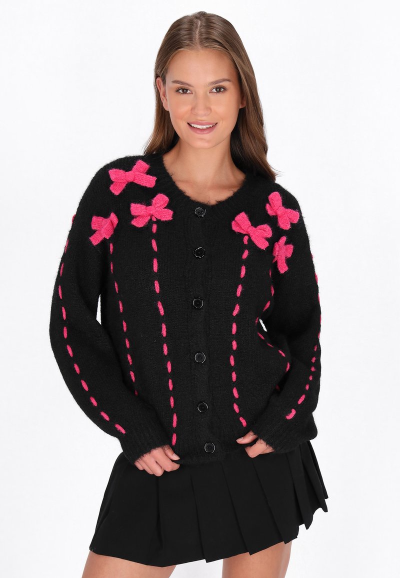 Juodas megztinis kardiganas su ryškiai rožinėmis kaspinais ir siūlėmis, mygtukų užsegimu, laisvu siluetu ir raukinėtomis rankogaliais. Suderintas su juoda klostuota sijonu.