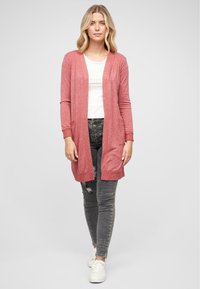 Long gilet rose clair avec ouverture sur le devant, deux poches avant, manches longues et poignets côtelés, associé à un haut blanc et un jean gris déchiré.