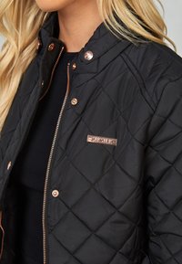 Chaqueta negra acolchada con herrajes y acentos en oro rosa, que presenta un cierre a presión y un parche con el logo en el lado izquierdo del pecho.