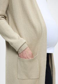 Beżowy kardigan wykonany z miękkiej dzianiny, z długimi rękawami, ściągaczami przy mankietach oraz przednią kieszenią. Noszony na białej bluzce.