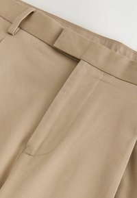Gros plan sur la ceinture d'un pantalon beige avec passants de ceinture et une braguette frontale dissimulée.