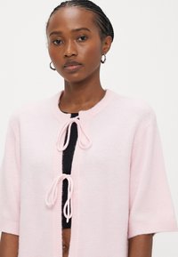 Light pink short-sleeve cardigan with a round neckline, featuring a front tie closure and ribbed accents. Soft, textured fabric.