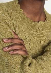 Person wearing a textured, olive green knitted sweater with scalloped neckline and fabric-covered buttons, arms crossed over chest.