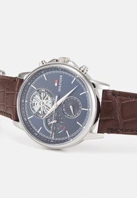 Reloj cronógrafo en tono plateado con esfera azul marino, correa de cuero marrón con patrón de cocodrilo y subesferas para la visualización de la fecha y la hora.