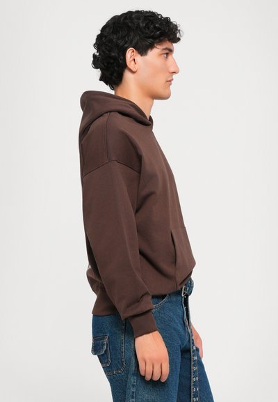 Giovane uomo con felpa marrone e jeans blu in piedi di profilo contro uno sfondo bianco uniforme.