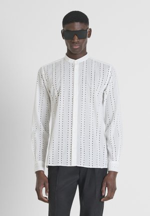 Camisa blanca de manga larga con patrones recortados verticales, cuello mandarín, cierre de botones, combinada con pantalones oscuros y gafas de sol negras.