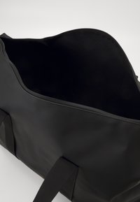 Sac de sport noir en matériau imperméable, doté d'une fermeture éclair, d'une texture douce et de poignées en tissu robustes. Intérieur ouvert sans compartiments.
