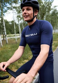Maillot de cyclisme bleu marine à manches courtes, avec fermeture éclair à l'avant et logo Castelli. Porté avec un casque noir tout en faisant du vélo.