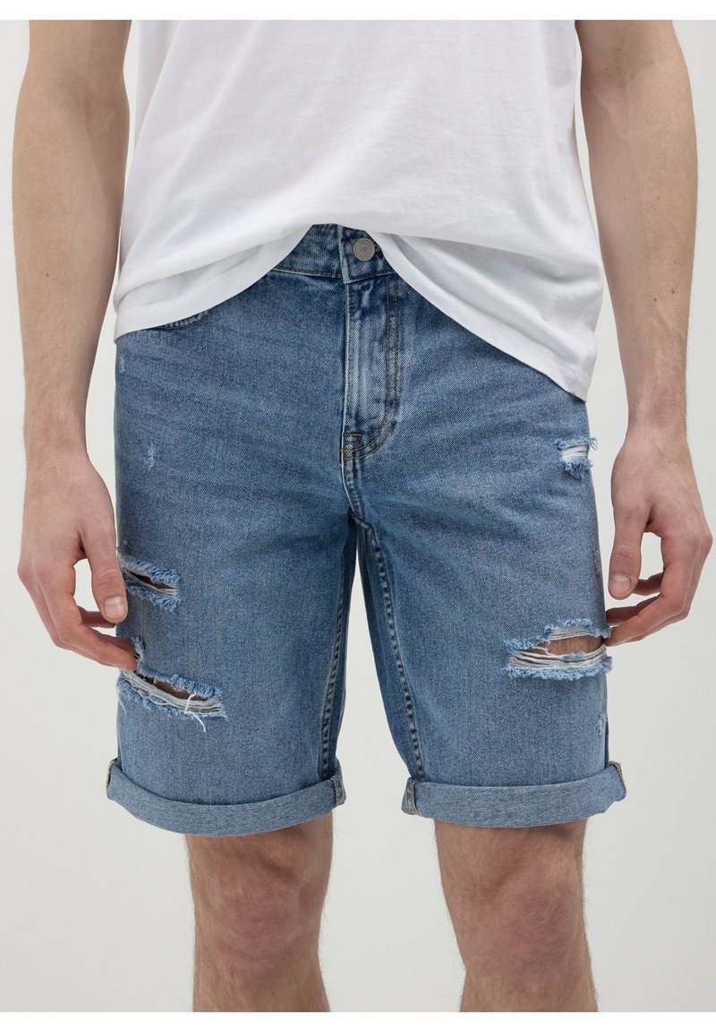 Homme portant un short en jean bleu usé et retroussé et un t-shirt blanc, debout avec les mains détendues le long du corps.