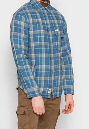 Homme portant une chemise à carreaux bleu et beige à boutons et un pantalon cargo marron, debout devant un fond uni.