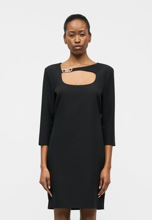 Femme portant une robe noire mi-longue aux manches trois-quarts avec une découpe carrée au niveau du décolleté, agrémentée d'une boucle argentée.