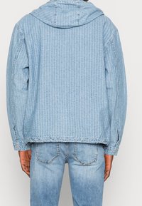 Ljusblå jeansjacka med en texturerad mönster och huva. Har en avslappnad passform och knapplösningar vid ärmarna och midjan.