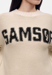 Femme portant un pull en tricot crème avec de grandes lettres noires cousues "SAMSON" sur la poitrine, montrant le cou et une partie du visage.