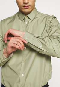 Camicia verde chiaro a maniche lunghe realizzata in tessuto morbido, con colletto a punta, chiusura con bottoni e polsini con un bottone e accenti dorati.