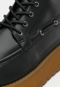 Chaussure en cuir noir avec lacets noirs passant par des œillets métalliques et semelle épaisse en caoutchouc brun à bande de roulement texturée.