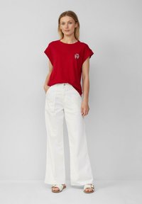 Vrouw in een rode top met korte mouwen met een klein krabontwerp, witte wijd uitlopende broek en witte sandalen, staand tegen een effen grijze achtergrond.