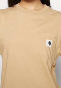 Beige t-shirt gjord av mjuk bomull, med rund hals och en vänster bröstficka med en liten svartvit logotag.