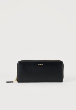 Portefeuille Coach en cuir noir avec fermeture éclair tout autour, logo doré et tirette de fermeture sur fond blanc.