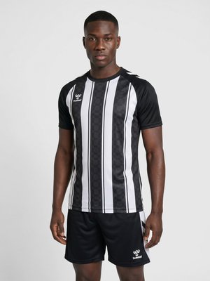 Man wearing a black and white striped sports jersey and black shorts standing against a plain light background.