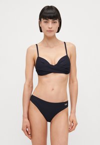 Črn bikini set, ki ima teksturirano tkanino, tanke naramnice, zvit dizajn na zgornjem delu in klasične spodnje dele z nizkim pasom. Opazen je tudi zlat nakit.