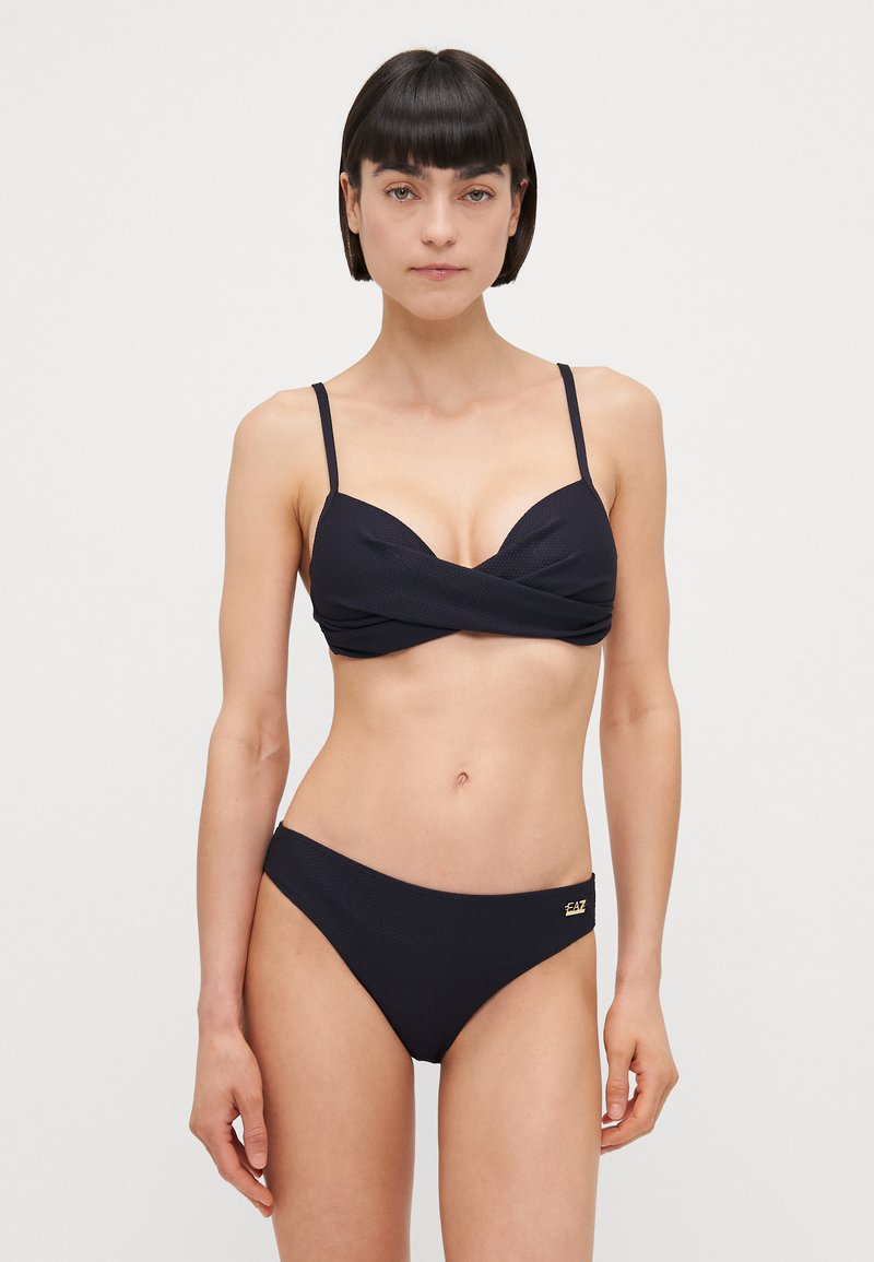 Črn bikini set, ki ima teksturirano tkanino, tanke naramnice, zvit dizajn na zgornjem delu in klasične spodnje dele z nizkim pasom. Opazen je tudi zlat nakit.