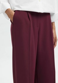 Personne portant un pantalon bordeaux ample à taille haute avec poches latérales, associé à une chemise blanche à manches longues rentrée dans le pantalon.