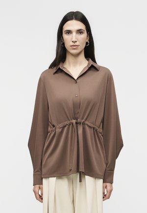 Γυναίκα με σκούρα μαλλιά φοράει καφέ πουκάμισο με κουμπιά και κορδόνι στη μέση, και εκρού παντελόνι με φαρδύ πόδι, στέκεται μπροστά από απλό λευκό φόντο.