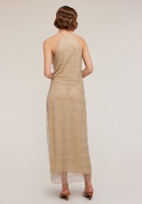 Beaded halter-neck dress in beige, featuring a sheer overlay with vertical, scalloped patterns and a fringed hem. Stylishly paired with heels.