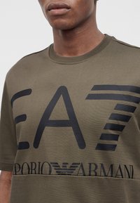 Νέος που φοράει ελαιοντυμένη μπλούζα Emporio Armani EA7 με μεγάλο μαύρο λογότυπο στο στήθος.