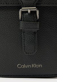 Fekete texturált bőrtáska részlet ezüst csattal és a pánt alatt dombornyomott "Calvin Klein" logóval.