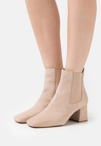 Botas de ante beiges hasta el tobillo con un tacón cuadrado y paneles laterales elásticos. Presentan una puntera afilada y una textura suave.