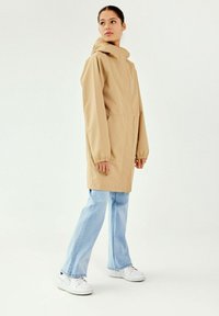 Veste imperméable beige avec une capuche, des poches avant et des poignets élastiques, assortie à un jean évasé bleu clair et des baskets blanches.