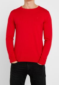 Pull-over en tricot rouge avec manches longues et col rond. Doté de poignets et d'un ourlet côtelés. Porté avec un jean noir.