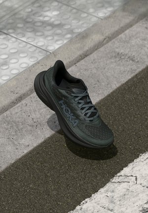 Czarny but biegowy z siateczkową cholewką, teksturowanymi detalami i logo, wyposażony w wyściełaną podeszwę i szare sznurówki, umieszczony na chodniku.