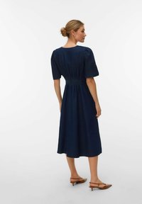 Kvinne iført en marineblå midi-kjole med puffede ermer og rynket midje, iført brune lave hæler, sett fra siden mot en hvit bakgrunn.