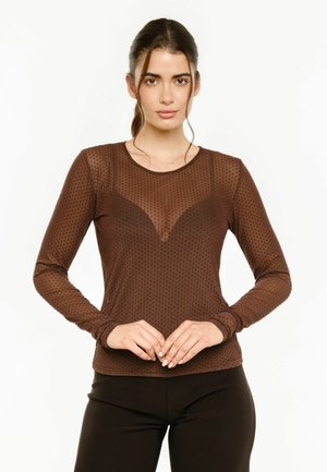 Femme aux cheveux foncés en queue de cheval portant un haut marron transparent à manches longues à pois et un pantalon noir, debout devant un fond blanc.