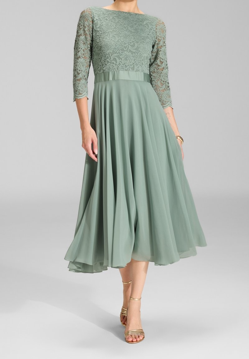 Femme portant une robe vert sauge avec corsage en dentelle et manches trois-quarts, jupe fluide et sandales dorées à talons hauts avec lanières.