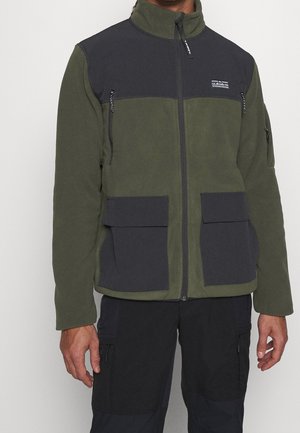 Mężczyzna w oliwkowo-zielonej i czarnej polarowej kurtce zapinanej na zamek, z wieloma kieszeniami na zamek i patki, w połączeniu z czarnymi spodniami cargo.