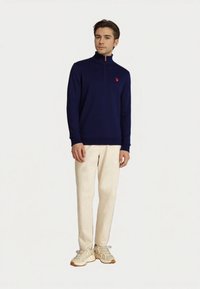 Junger Mann in einem navyfarbenen Quarter-Zip-Pullover, beigen Hosen und beigen Turnschuhen, der vor einem schlichten weißen Hintergrund steht.