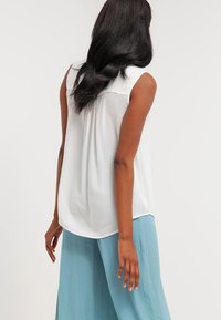 White sleeveless blouse made of lightweight material, featuring a rounded hem and subtle texture. Paired with light blue flowy pants.