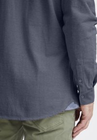Camicia grigia a maniche lunghe in tessuto texturizzato, con polsini bottonati e orlo curvo, abbinata a pantaloni verdi oliva. Dettaglio delle cuciture visibile.