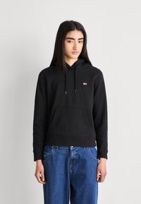 Sudadera negra con capucha, bolsillo frontal, cordón y un pequeño logo en el pecho. Presenta un ajuste relajado con puños de canalé.