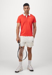 Uomo che indossa una polo rossa e pantaloni corti bianchi, in piedi contro uno sfondo bianco uniforme, mentre tiene in mano una racchetta da tennis.