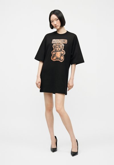Γυναίκα με κοντά μαύρα μαλλιά που φοράει μαύρο αμάνικο φόρεμα oversized με πορτοκαλί αρκουδάκι και την επιγραφή "Moschino", συνδυασμένο με μαύρες γόβες.