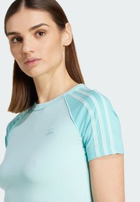 Svijetloplava sportska majica s kratkim rukavima, s tri bijele pruge na ramenima i suptilnim logotipom na prsima. Glatka tekstura tkanine.