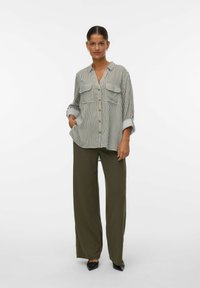 Une femme portant une chemise à rayures lâche avec des manches retroussées, un pantalon large olive et des chaussures noires à bout pointu, se tenant contre un fond blanc.