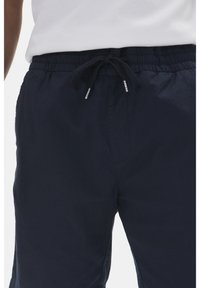 Personne portant un pantalon à cordon de serrage bleu marine et une chemise blanche, avec un focus sur la taille et la zone supérieure de la cuisse.